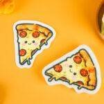 Pizza slice-shaped cellulose sponges with melted cheese illustration, photographed on warm yellow surface with cookies and bread decor.