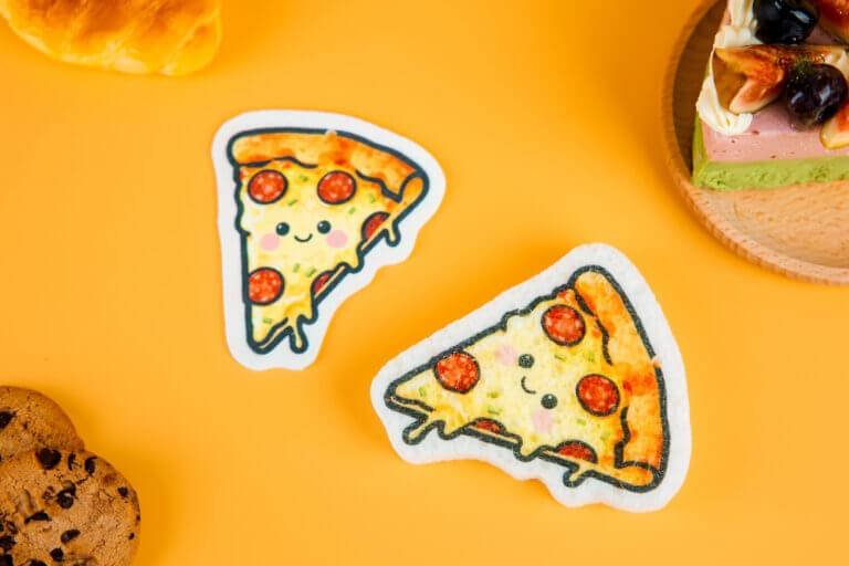 Pizza slice-shaped cellulose sponges with melted cheese illustration, photographed on warm yellow surface with cookies and bread decor.