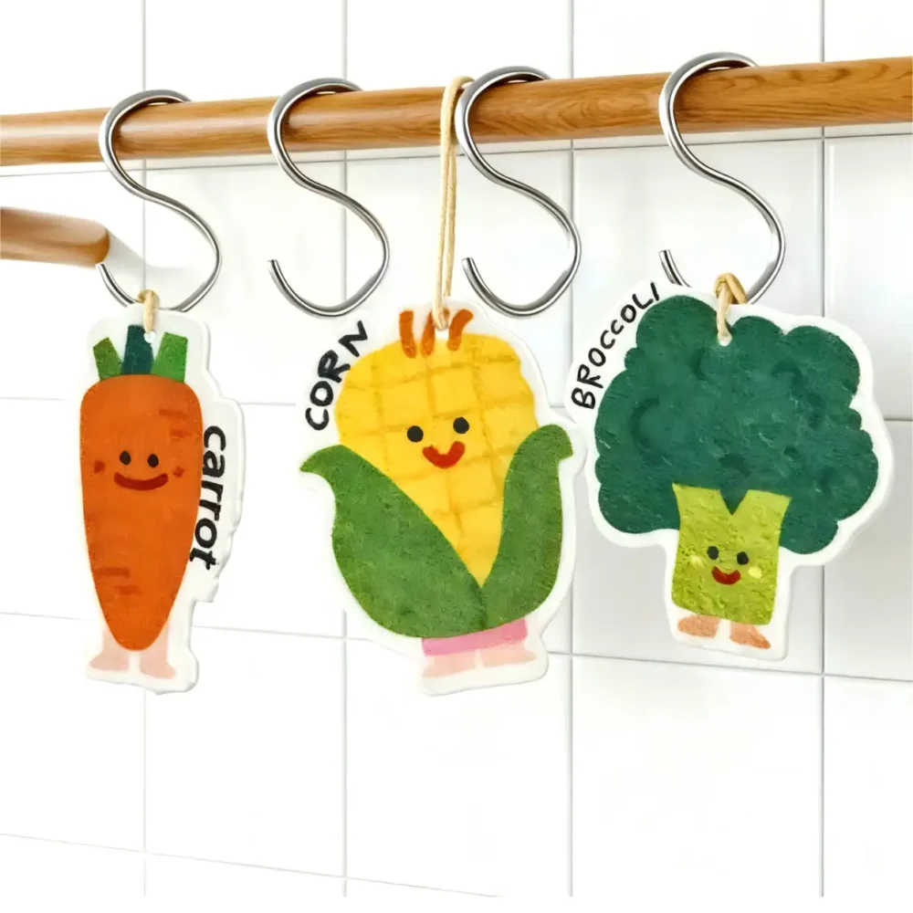 Carrot, corn, and broccoli cellulose sponges hanging from white hooks by their cords to dry.